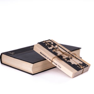 May include: A wooden bookmark with a black cutout design of a tree and the text "Reading is good for you". The bookmark is tied with twine and is resting on top of a black hardcover book.
