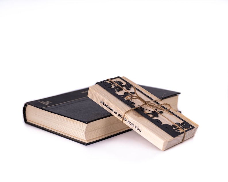 May include: A wooden bookmark with a black cutout design of a tree and the text "Reading is good for you". The bookmark is tied with twine and is resting on top of a black hardcover book.