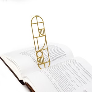 Gold metal bookmark with a modern Bauhaus geometric design standing upright in the pages of an open book — sleek and artistic reading accessory.