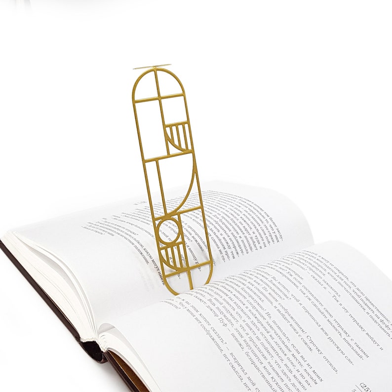 Gold metal bookmark with a modern Bauhaus geometric design standing upright in the pages of an open book — sleek and artistic reading accessory.