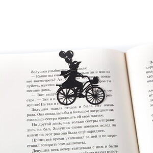 Black metal bike bookmark with a girl riding a bicycle holding balloons, placed inside an open book on a white background.