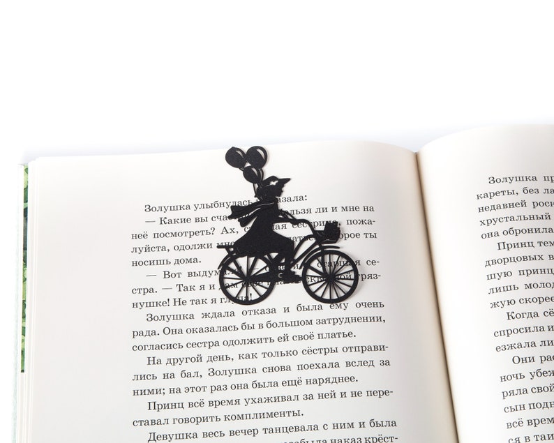 Black metal bike bookmark with a girl riding a bicycle holding balloons, placed inside an open book on a white background.