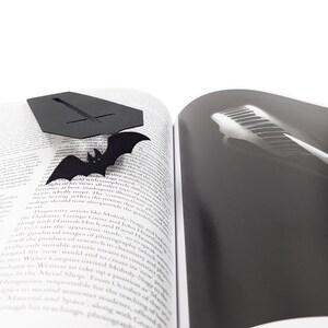 Black metal coffin-shaped bookmark with a flying bat detail resting on the open pages of a book; one page shows text and the other shows a black-and-white photo of piano keys.