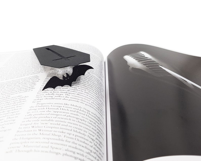 Black metal coffin-shaped bookmark with a flying bat detail resting on the open pages of a book; one page shows text and the other shows a black-and-white photo of piano keys.