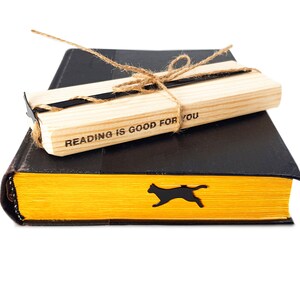 Black metal cat bookmark shaped like a running cat resting inside a closed yellow-edged book, paired with a wooden block engraved “Reading is good for you.”