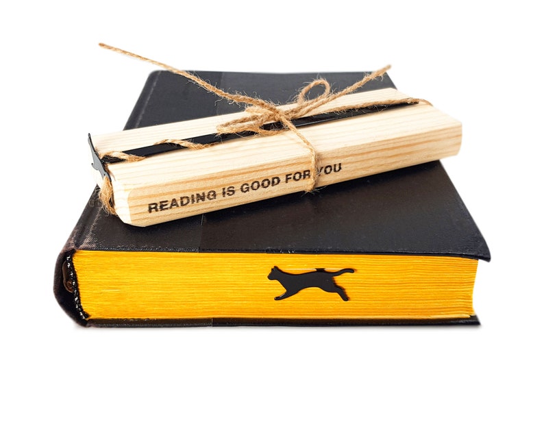 Black metal cat bookmark shaped like a running cat resting inside a closed yellow-edged book, paired with a wooden block engraved “Reading is good for you.”