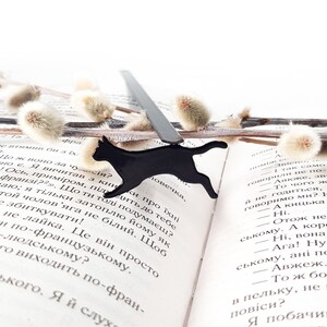 Black metal running cat bookmark resting on an open book with text in Ukrainian, surrounded by soft pussy willow branches.