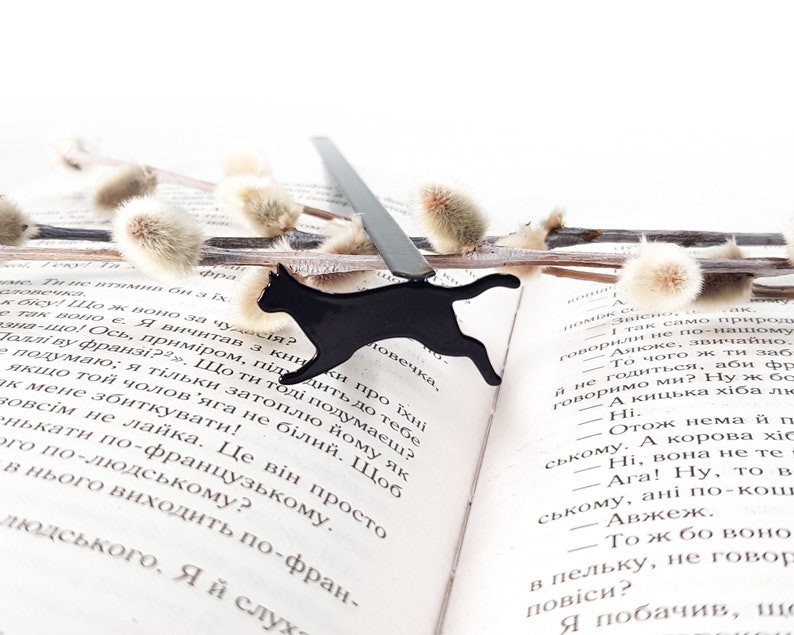 Black metal running cat bookmark resting on an open book with text in Ukrainian, surrounded by soft pussy willow branches.