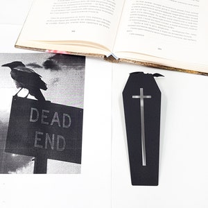 Black metal coffin-shaped bookmark with a cutout cross and small bat detail placed on a white surface beside an open book and a black-and-white print of a crow sitting on a “DEAD END” sign.