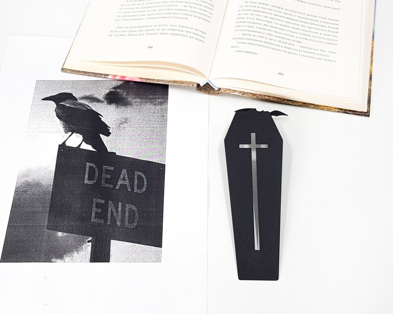 Black metal coffin-shaped bookmark with a cutout cross and small bat detail placed on a white surface beside an open book and a black-and-white print of a crow sitting on a “DEAD END” sign.