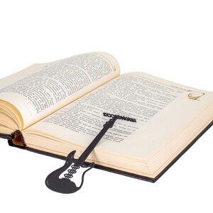 Black metal bookmark shaped like an electric guitar with READING ROCKS along the neck, placed inside an open book. A bold, fun gift for music-loving readers.