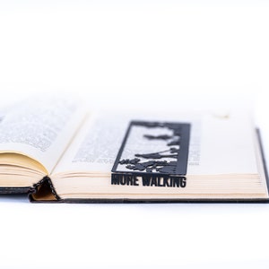 May include: A black metal bookmark with a silhouette of people walking and the text "MORE WALKING". The bookmark is placed in an open book.