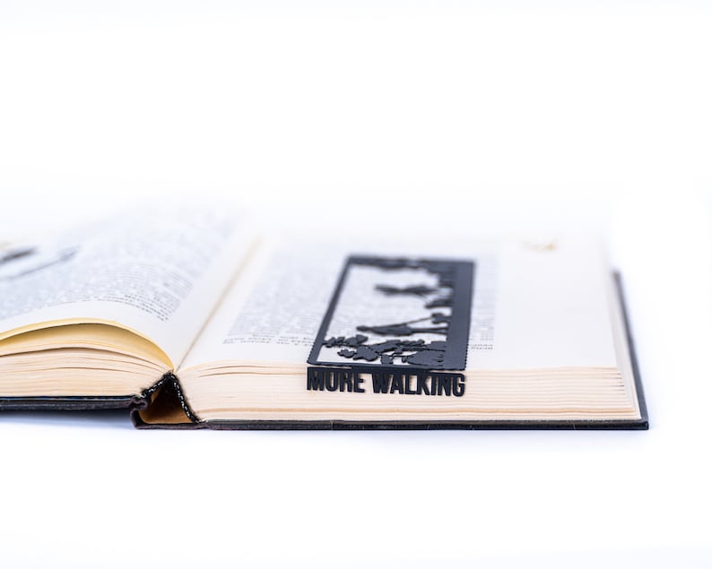 May include: A black metal bookmark with a silhouette of people walking and the text "MORE WALKING". The bookmark is placed in an open book.