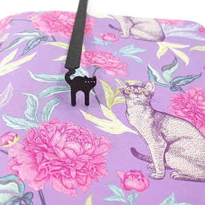 May include: A black cat-shaped bookmark hangs over a book with a floral pattern. The book's pages display a purple background with pink peonies and illustrations of cats. The bookmark is made of a dark material and has a simple, stylized design.