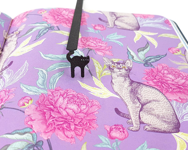 May include: A black cat-shaped bookmark hangs over a book with a floral pattern. The book's pages display a purple background with pink peonies and illustrations of cats. The bookmark is made of a dark material and has a simple, stylized design.