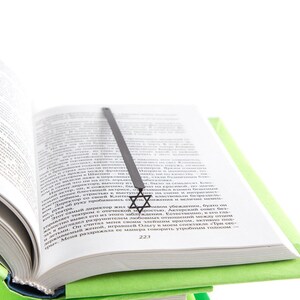 Black metal Star of David bookmark resting in an open book, placed on a green hardcover, minimalist and symbolic design.