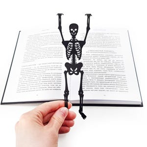 A hand holding a black metal skeleton-shaped bookmark in front of an open book, the skeleton’s arms raised as if climbing out of the pages.