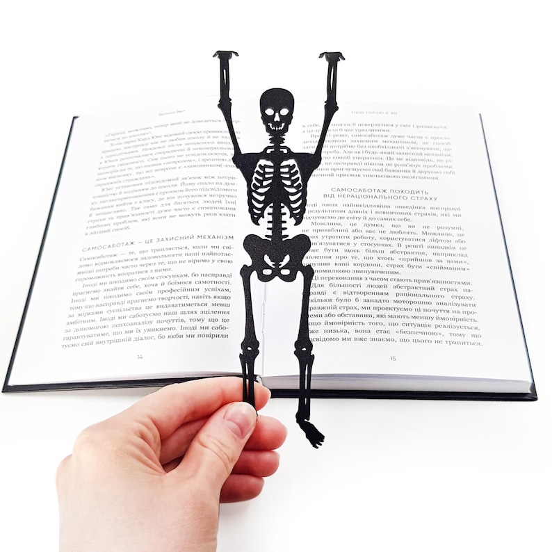 A hand holding a black metal skeleton-shaped bookmark in front of an open book, the skeleton’s arms raised as if climbing out of the pages.