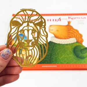 May include: A gold-coloured metal bookmark shaped like a lion's head. The bookmark is detailed with intricate cutouts and has a shiny finish.