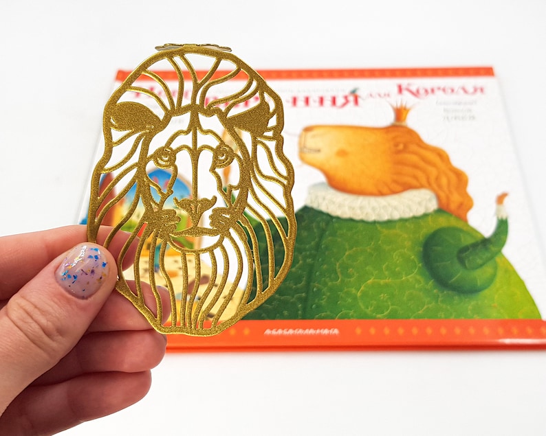 May include: A gold-coloured metal bookmark shaped like a lion's head. The bookmark is detailed with intricate cutouts and has a shiny finish.