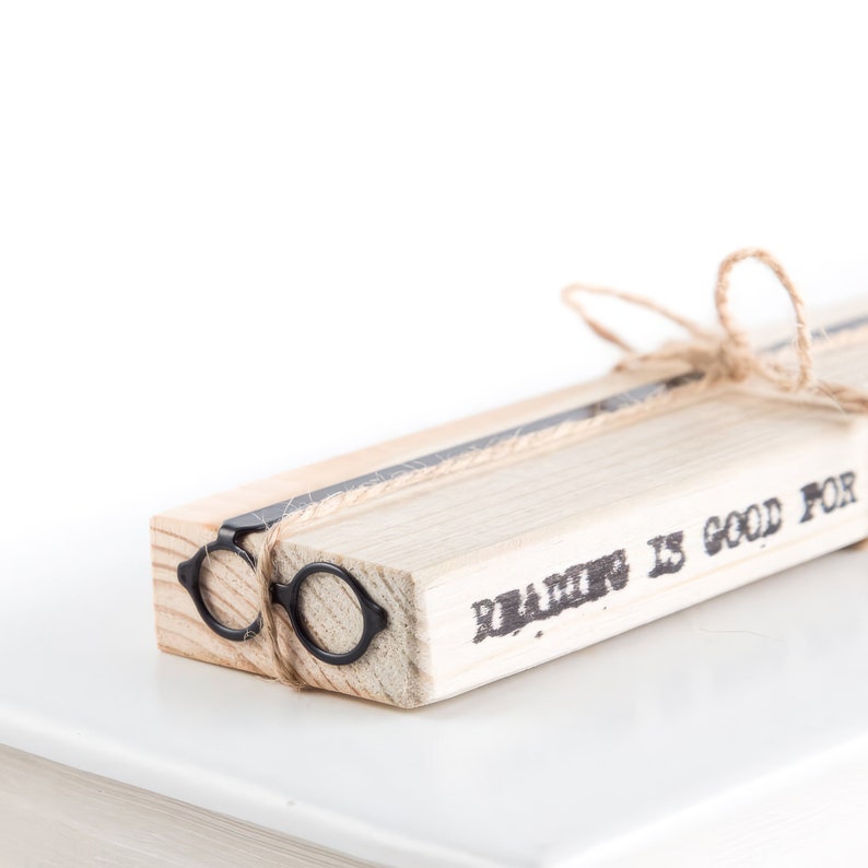 Black metal page saver shaped like round architect's glasses, tied to a wooden block with the phrase ‘Reading is good for you’ printed on the side.
