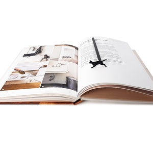 Black metal running cat bookmark resting across the open pages of a photo book featuring minimalist interior designs.
