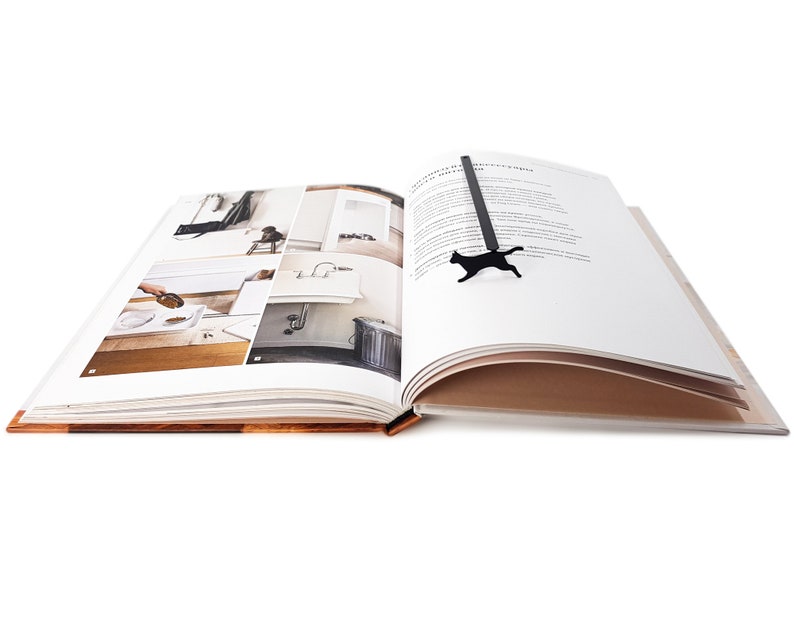 Black metal running cat bookmark resting across the open pages of a photo book featuring minimalist interior designs.