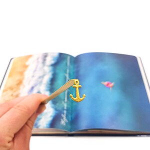 May include: A gold anchor bookmark is placed in a book with a watercolor image of a beach and ocean waves. The bookmark is made of metal and has a shiny finish.