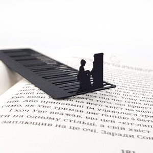 May include: Black metal bookmark featuring a piano keyboard design with a silhouette of a person playing. The bookmark is placed on the open pages of a book, adding a touch of elegance to reading.