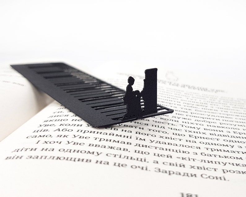 May include: Black metal bookmark featuring a piano keyboard design with a silhouette of a person playing. The bookmark is placed on the open pages of a book, adding a touch of elegance to reading.