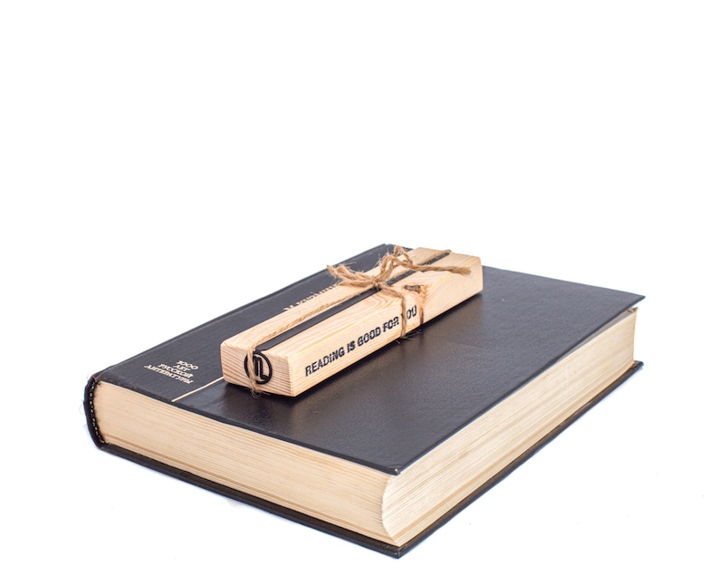 May include: A wooden bookmark with the text "Reading is good for you" tied with twine, resting on top of a closed black hardcover book.