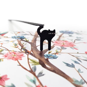 May include: A black cat-shaped bookmark with a long, thin arm, perched on a floral-patterned page. The page features a watercolor design of a tree with pink and blue flowers and a bird.