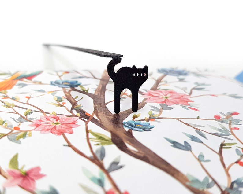 May include: A black cat-shaped bookmark with a long, thin arm, perched on a floral-patterned page. The page features a watercolor design of a tree with pink and blue flowers and a bird.
