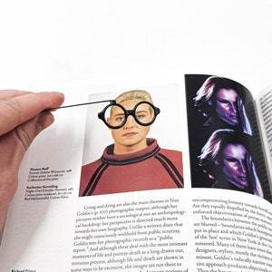 Black metal page saver shaped like architect glasses held over a fashion magazine portrait, adding a modernist twist to the model’s face. Minimalist and clever reading accessory.