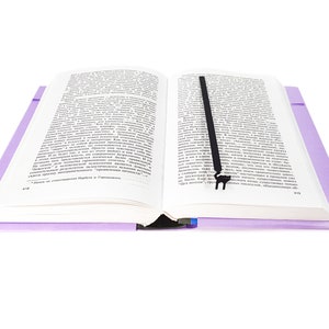 May include: An open book with text, featuring a black bookmark. The book has a light purple cover and is set against a white background. The bookmark is a simple, elegant design.