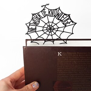 May include: A black paper cut-out bookmark shaped like a spider web with the words "Network of Knowledge" around the edge. The bookmark is placed on top of a brown book.