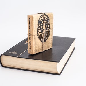 May include: A wooden book with the text "Reading is good for you" and a lion silhouette on the cover. The book is sitting on top of a black hardback book.