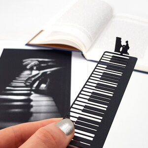 May include: A black metal bookmark shaped like a piano keyboard, with a silhouette of a person playing the piano at the top. The bookmark is held in front of an open book and a black and white photo of piano keys.