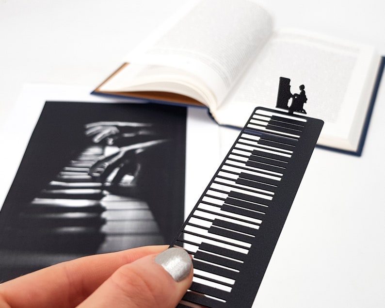 May include: A black metal bookmark shaped like a piano keyboard, with a silhouette of a person playing the piano at the top. The bookmark is held in front of an open book and a black and white photo of piano keys.