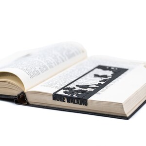 May include: A black metal bookmark with a silhouette of people walking and the text "MUME WALKING". The bookmark is placed in an open book.