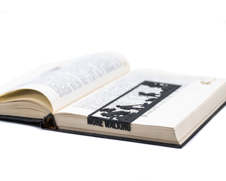 May include: A black metal bookmark with a silhouette of people walking and the text "MUME WALKING". The bookmark is placed in an open book.