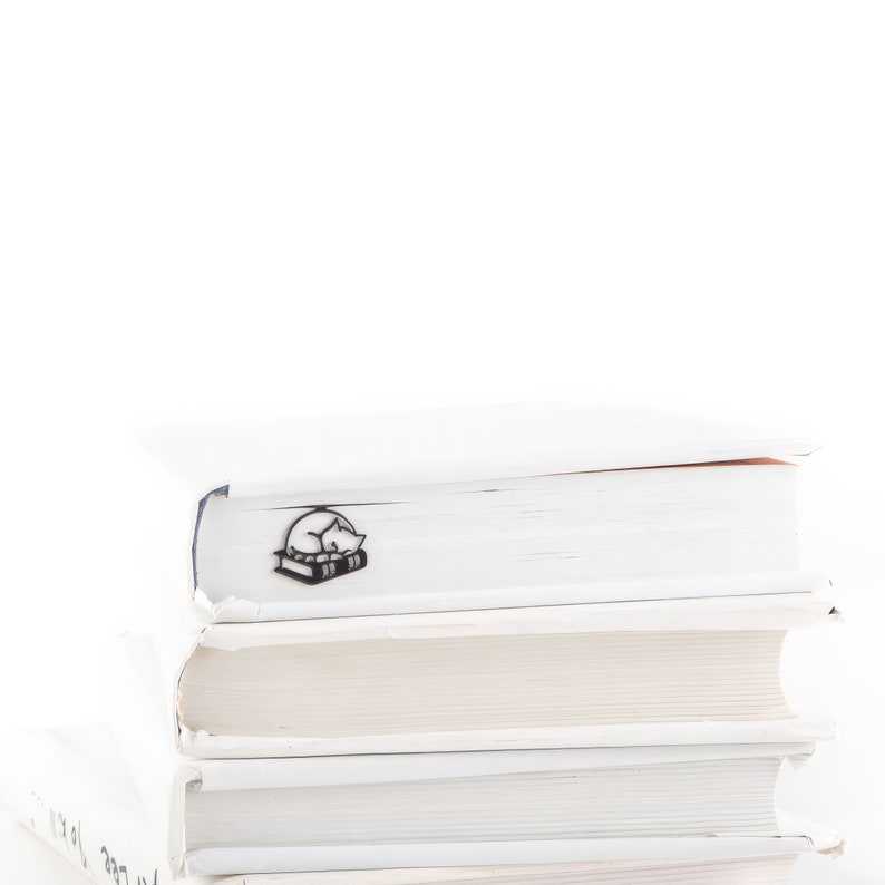 Op de afbeelding: Zwart metalen boekenlegger met een slapende kat ontwerp, geplaatst op een stapel witte boeken. De boekenlegger toont een kat die op een stapel boeken ligt. De boeken hebben witte kaften en zijn op elkaar gestapeld.