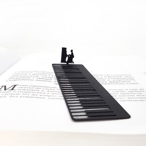 May include: A black metal bookmark shaped like a piano keyboard, with a silhouette of a person playing the piano at the top. The bookmark is placed in an open book, with the pages visible.