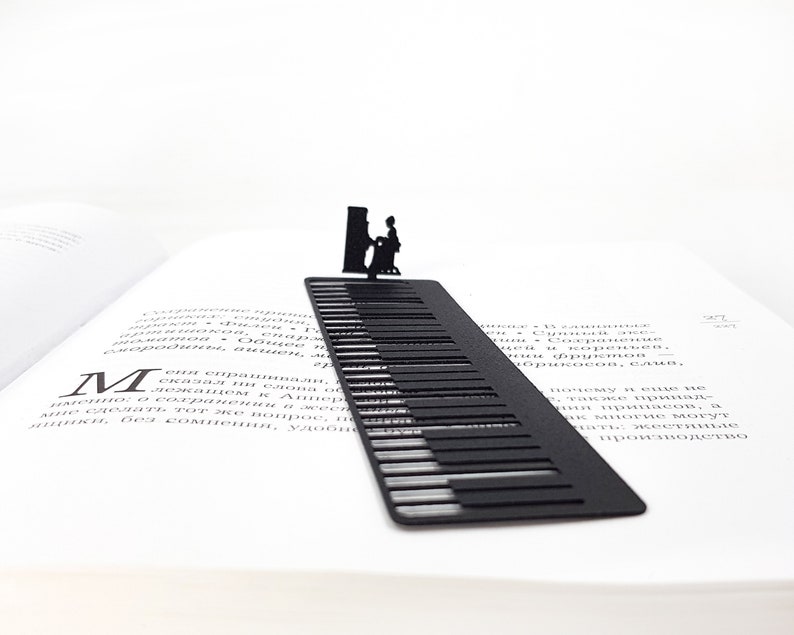 May include: A black metal bookmark shaped like a piano keyboard, with a silhouette of a person playing the piano at the top. The bookmark is placed in an open book, with the pages visible.