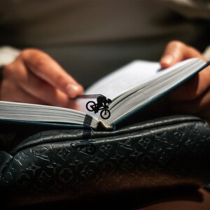 A black metal bookmark shaped like a person riding a bicycle. The bookmark is placed in a book with white pages.