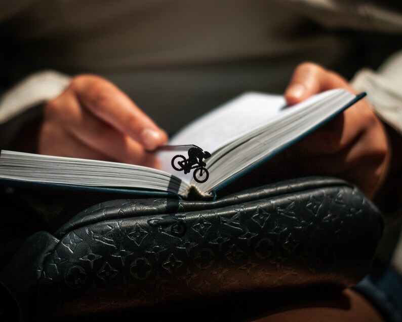 A black metal bookmark shaped like a person riding a bicycle. The bookmark is placed in a book with white pages.