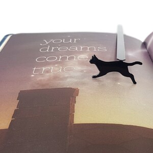 Black metal running cat bookmark resting on an open book with the text “your dreams come true” printed on a softly lit background.