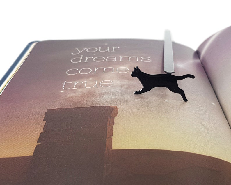 Black metal running cat bookmark resting on an open book with the text “your dreams come true” printed on a softly lit background.