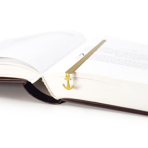 May include: A gold-colored metal bookmark shaped like an anchor. The bookmark is placed in a book with the pages open.