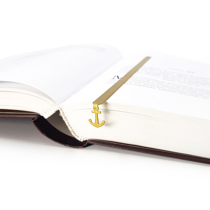 May include: A gold-colored metal bookmark shaped like an anchor. The bookmark is placed in a book with the pages open.